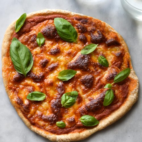 A gluten free sourdough margherita pizza against a white marble backdrop with a glass of water in the top right corner