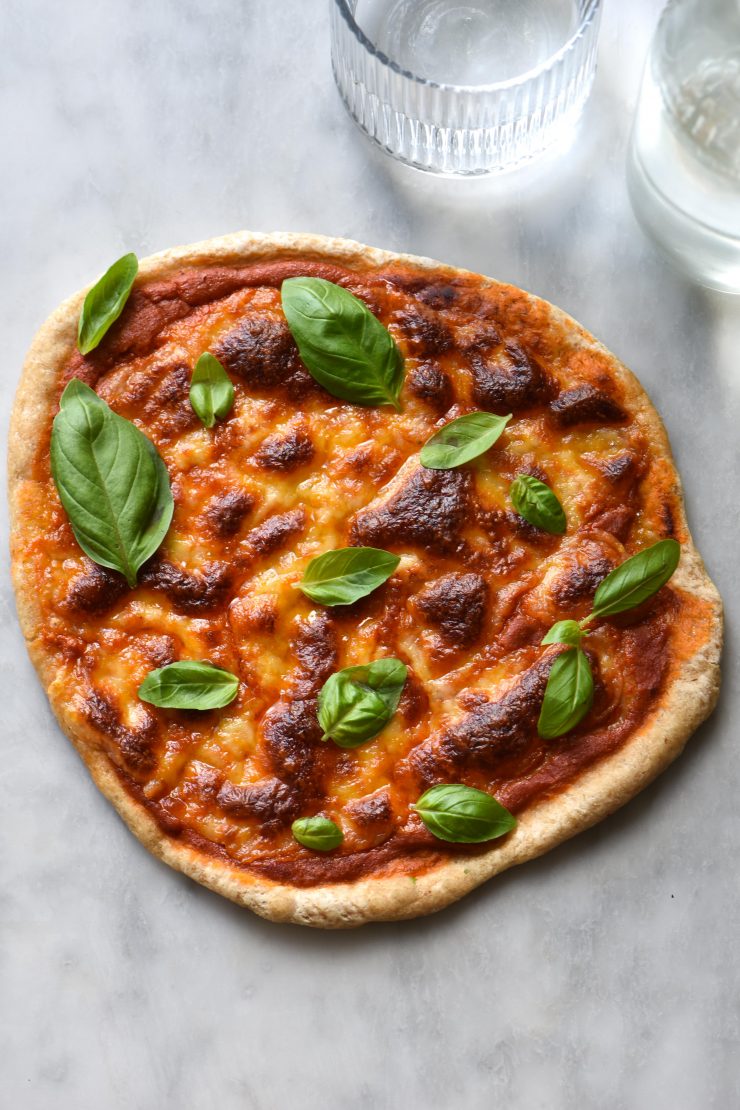 A gluten free sourdough margherita pizza against a white marble backdrop with a glass of water in the top right corner