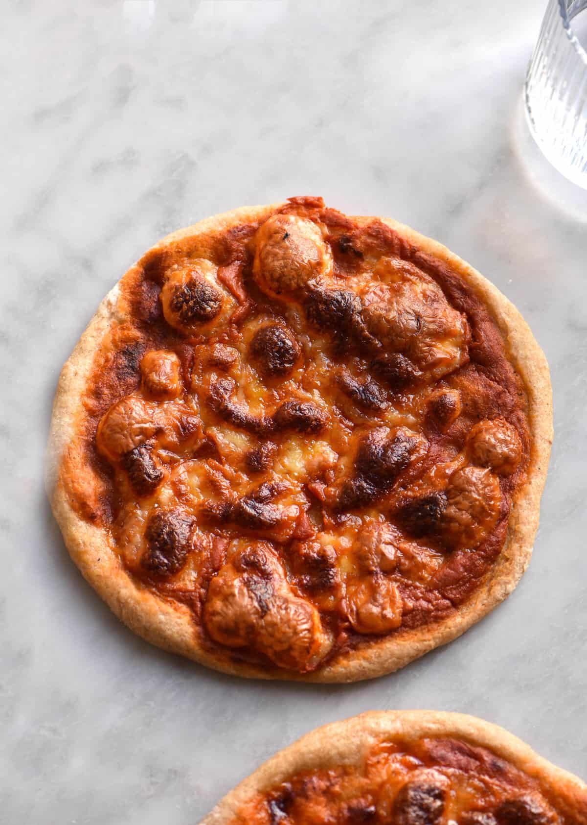 A close up of a gluten free sourdough pizza. The pizza is topped with margherita style toppings - red pizza sauce and bubbly browned mozzarella. It is set against a white marble backdrop