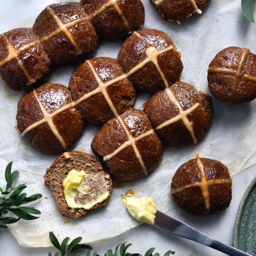 Gluten free, vegan and FODMAP friendly hot cross buns against a white marble backdrop surrounding by green florals