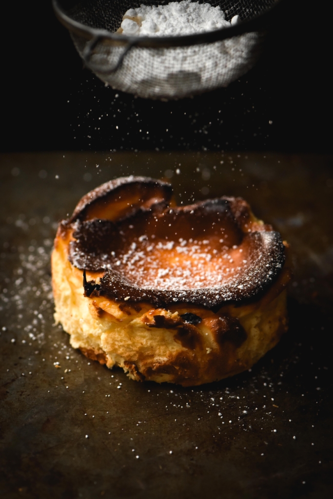 A side on view of a mini cheesecake being sprinkled with icing sugar against a dark metal backdrop