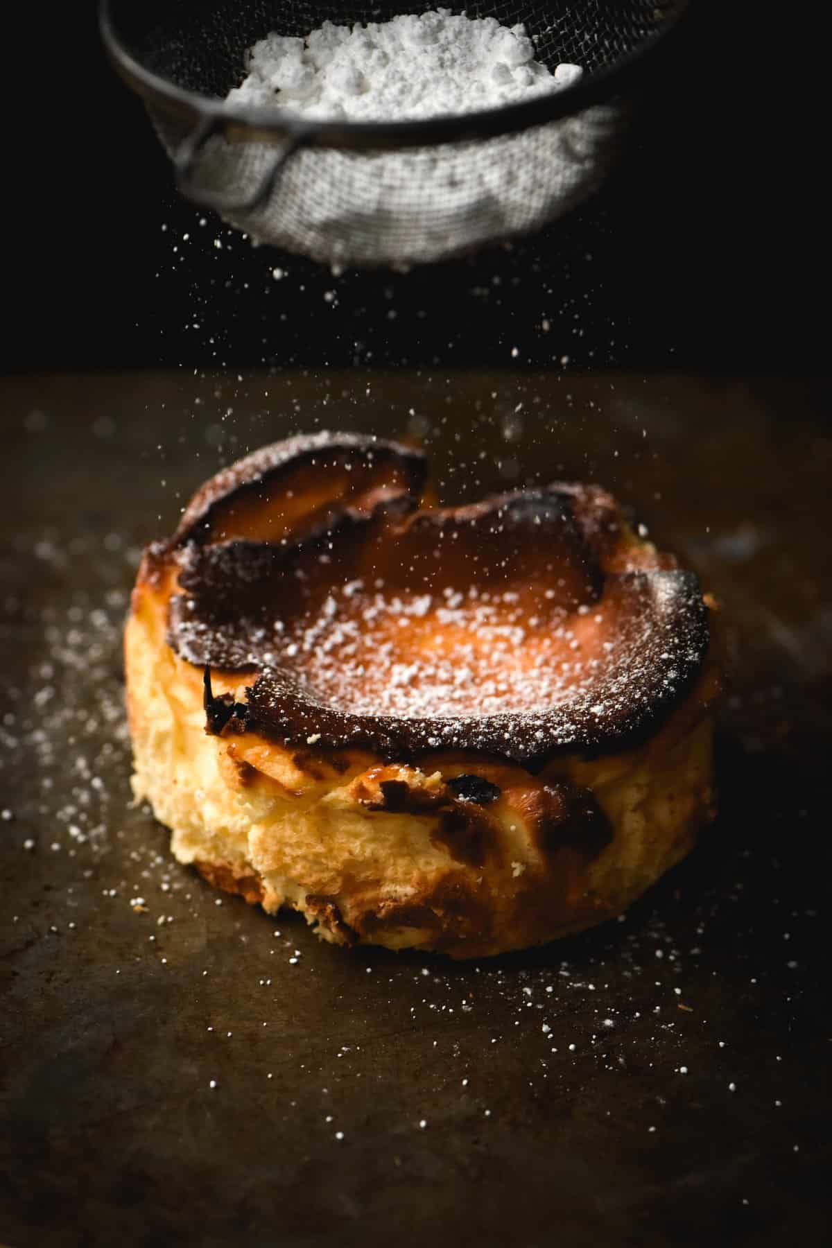 A side on view of a mini gluten free cheesecake being sprinkled with icing sugar against a dark metal backdrop