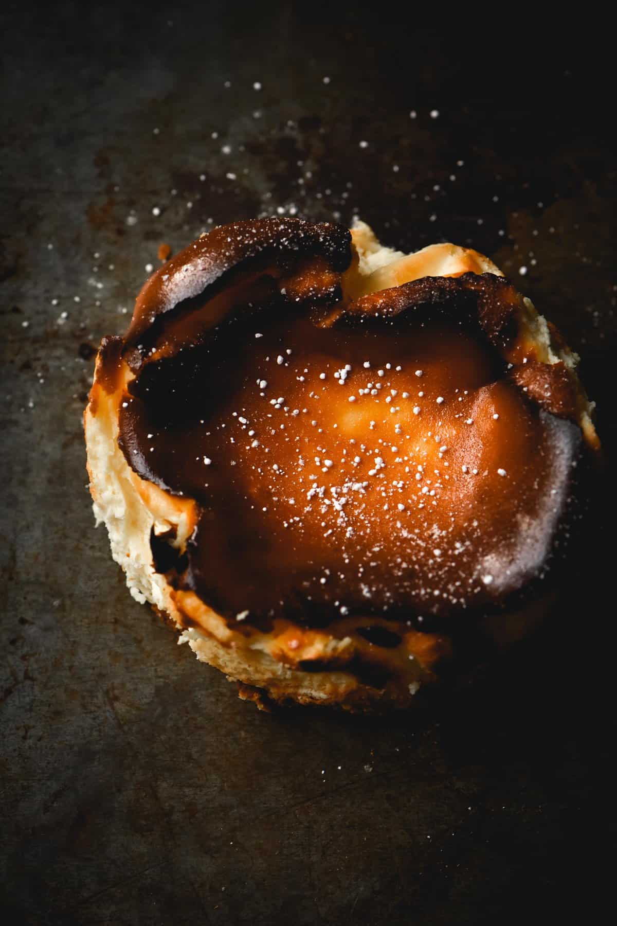 A moody aerial view of a Burnt Basque style mini cheesecake, sprinkled with icing sugar and set against a dark metal backdrop