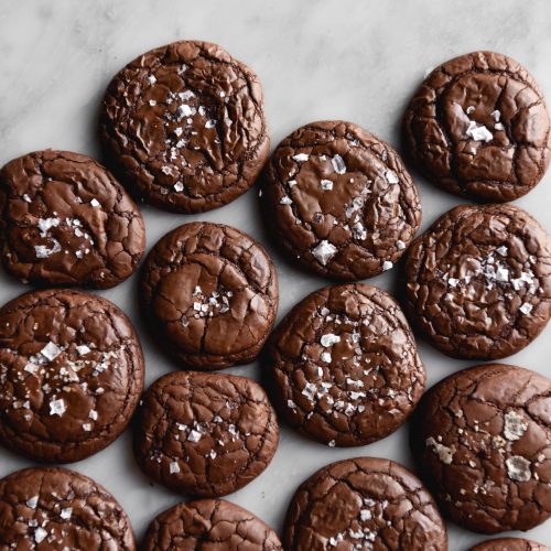 An aerial view of gluten free brownie cookies arranged on a white marble table. The cookies are crinkly and topped with sea salt flakes