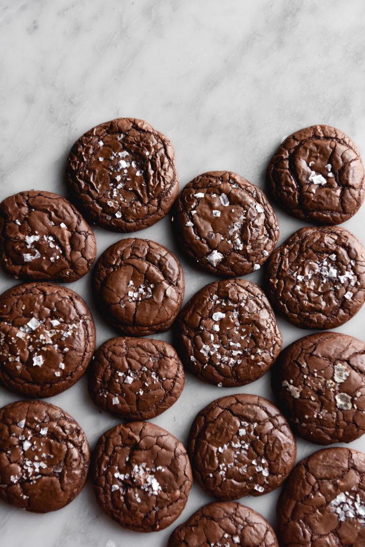 An aerial view of gluten free brownie cookies arranged on a white marble table. The cookies are crinkly and topped with sea salt flakes