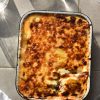 A tray of FODMAP friendly vegetarian moussaka sitting on a light tiled surface in bright sunlight. The top of the moussaka is cheesy, golden and caramelised. Around the edges of the moussaka tray and glasses of water lit with sunlight, a plate and a white linen cloth.