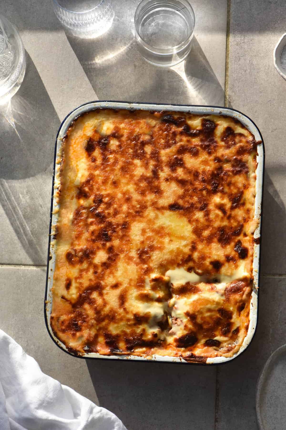 A tray of FODMAP friendly vegetarian moussaka sitting on a light tiled surface in bright sunlight. The top of the moussaka is cheesy, golden and caramelised. Around the edges of the moussaka tray and glasses of water lit with sunlight, a plate and a white linen cloth.