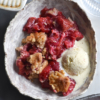 An aerial shot of a bowl of gluten free fruit crumble, made with berries. A scoop of melting vanilla ice cream sits alongside the crumble in a white ceramic bowl atop a white marble table. Two other plates of crumble surround the central bowl