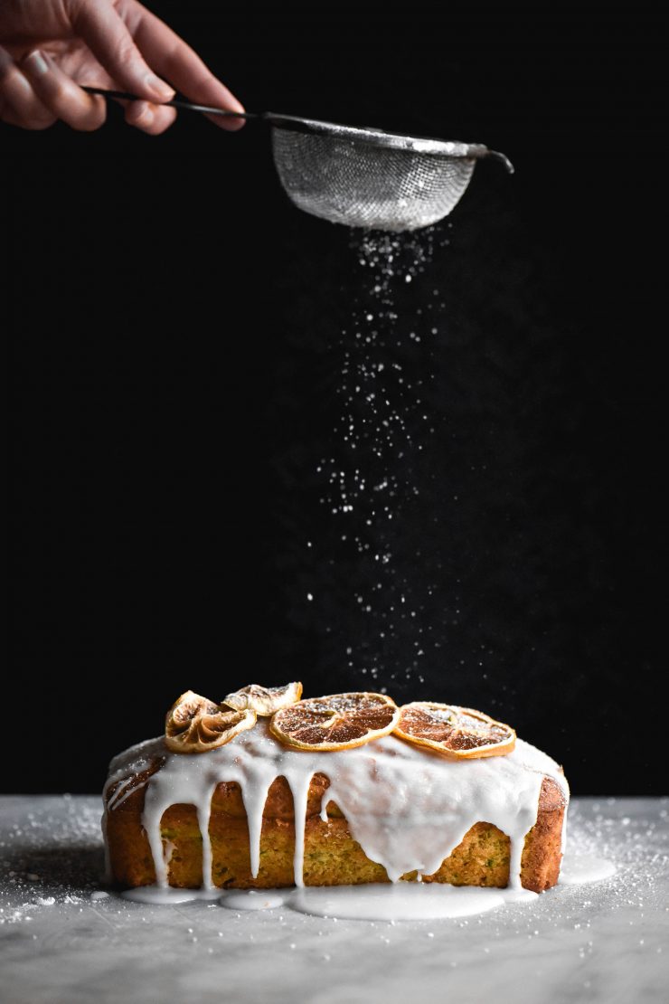 A side on view of a gluten free zucchini loaf set against a dark backdrop. The loaf is adorned with drizzled icing and dehydrated lemon. A dusting of icing sugar falls onto the loaf from a hand holding a sieve at the top of the image.