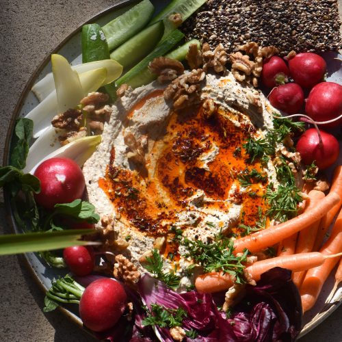 An aerial view of FODMAP friendly hummus platter surrounded by vibrant vegetable crudités. The hummus is drizzled with a red chilli oil, and the platter is set against stone tile, dappled in sunglight.
