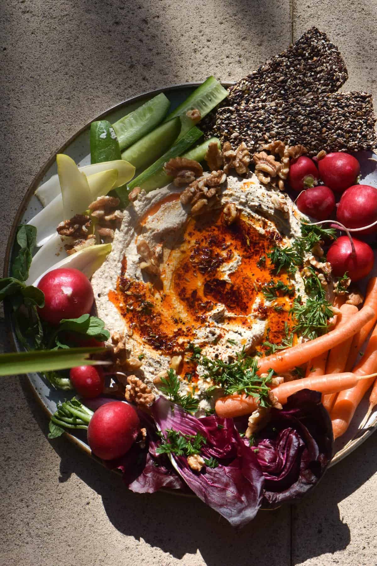 An aerial view of FODMAP friendly hummus platter surrounded by vibrant vegetable crudités. The hummus is drizzled with a red chilli oil, and the platter is set against stone tile, dappled in sunglight.