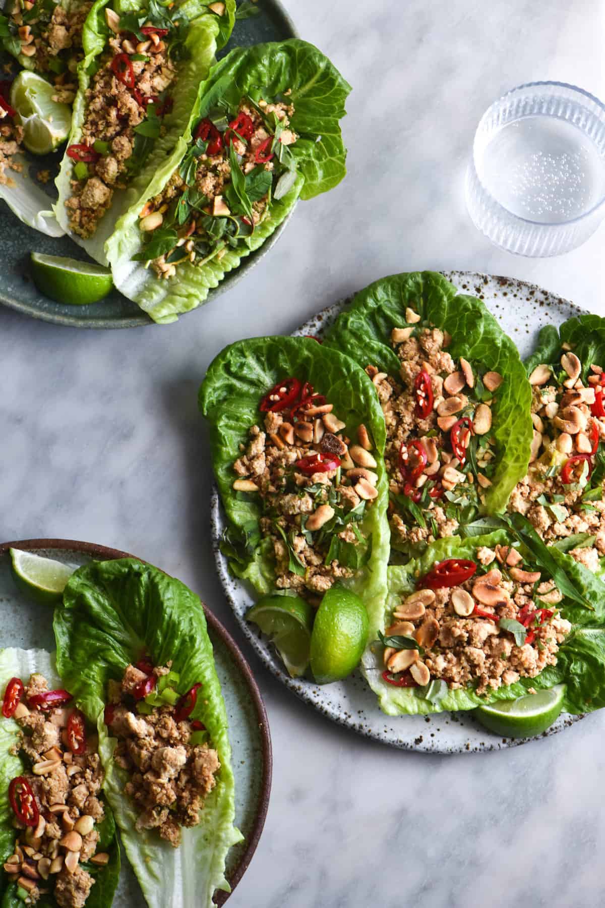 An aerial view of vegan tofu larb lettuce cups sitting atop a white marble table. The larb lettuce cups are casually arranged and topped with fresh sliced chillies, toasted peanuts and fresh herbs