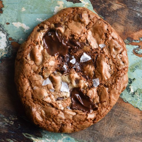 A close up of a gluten free tahini cookie studded with melting dark chocolate and topped with sea salt flakes. It is set against a worn wooden backdrop with grain and white paint marks