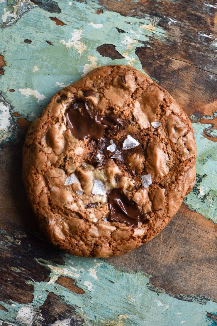 A close up of a gluten free tahini cookie studded with melting dark chocolate and topped with sea salt flakes. It is set against a worn wooden backdrop with grain and white paint marks