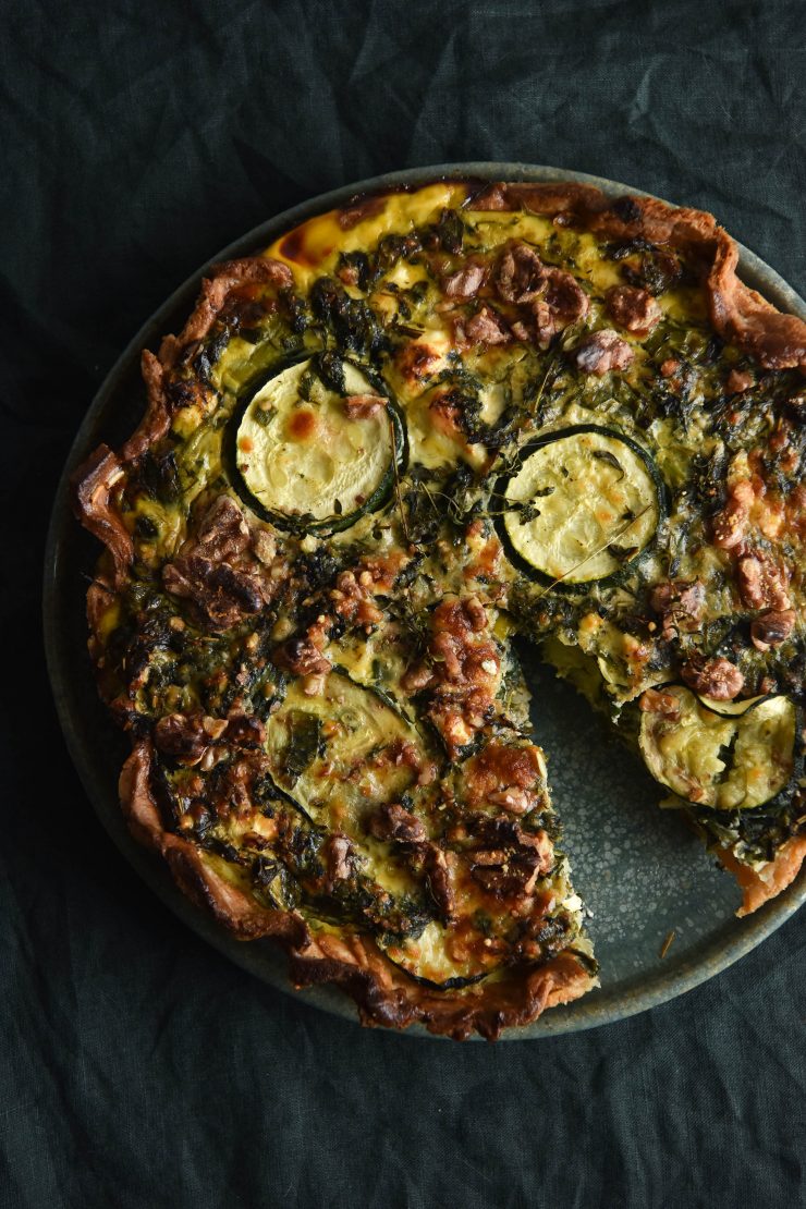 A close up shot of a gluten free spanakopita tart topped with zucchini coins and crushed walnuts. A slice of the tart has been removed, revealing a green ceramic plate underneath. The scene is set against an olive linen green tablecloth