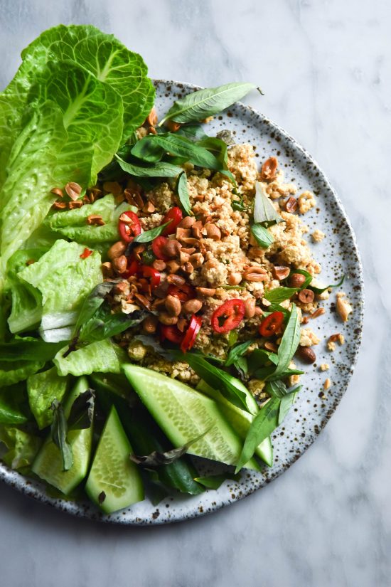 A white ceramic speckled plate of vegan tofu larb set against a white marble backdrop. The plate is bursting with tofu topped with freshly chopped chilli and toasted peanuts, alongside verdant green lettuce, cucumber wedges and fresh herbs.