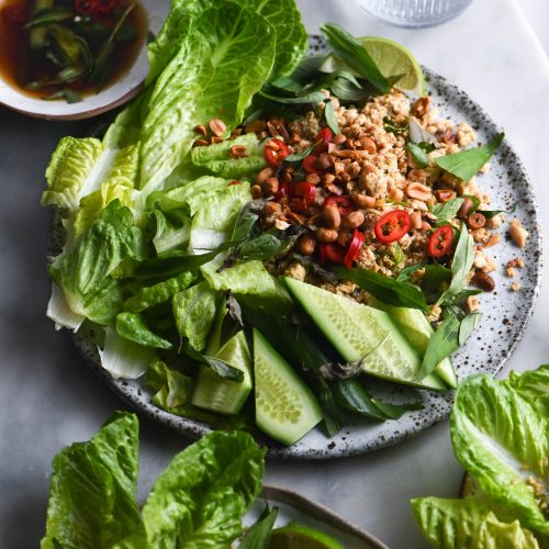 Three plates of vegan tofu larb sitting atop a marble table. The plates are casually arranged with tofu larb, topped with fresh chillies and toasted peanuts, as well as lots of fresh lettuce, cucumber and herbs. A bowl of vegan nuoc mam sits in the back left corner of the image.