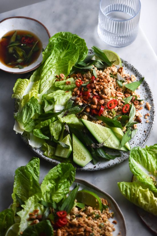 Three plates of vegan tofu larb sitting atop a marble table. The plates are casually arranged with tofu larb, topped with fresh chillies and toasted peanuts, as well as lots of fresh lettuce, cucumber and herbs. A bowl of vegan nuoc mam sits in the back left corner of the image.
