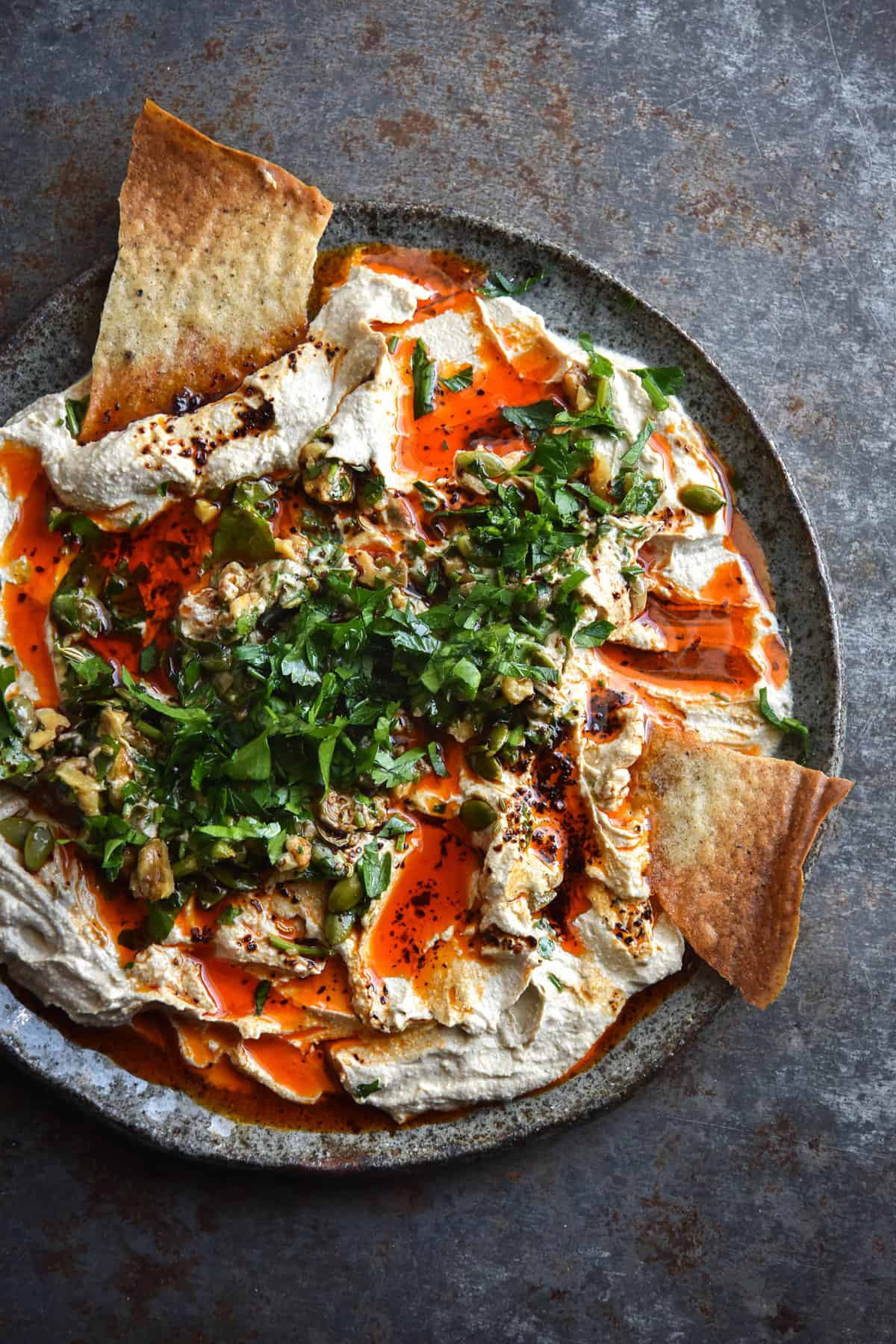 A messy plate of FODMAP friendly hummus topped with a walnut gremolata and vibrant red chilli oil. The plate of hummus is messy and rustic, with a number of chip indents in the dip. It is set against a grey steel backdrop and set on a grey ceramic plate.