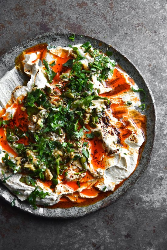 A messy plate of FODMAP friendly hummus topped with a walnut gremolata and vibrant red chilli oil. The plate of hummus is messy and rustic, with a number of chip indents in the dip. It is set against a grey steel backdrop and set on a grey ceramic plate.
