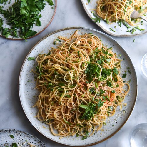 An aerial view of three white ceramic plates loaded with FODMAP friendly pasta aglio e olio. The spaghetti is topped with fresh chopped parsley, chilli flakes and finely grated parmesan. The plates are set against a white marble backdrop and a plate of extra chopped parsley sits in the top lefthand corner.