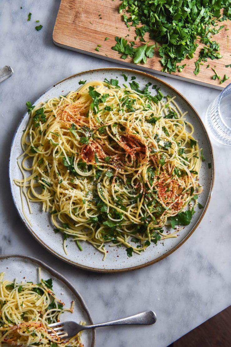 An aerial view of two plates of FODMAP friendly spaghetti aglio e olio. The spaghetti sits atop two white ceramic plates of varying sizes and is rustically presented. A chopping board topped with chopped parsley sits in the top righthand corner, and a fork pokes out from a plate of spaghetti in the bottom lefthand corner.