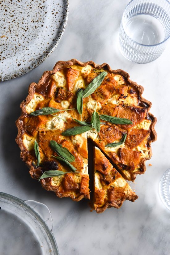 An aerial image of a gluten free pumpkin, sage and goats cheese tart sitting on a white marble backdrop. The tart is a deep orange, dotted with melted goats cheese and topped with fresh sage leaves. The tart is casually surrounded by water glasses, plates and an empty pie dish