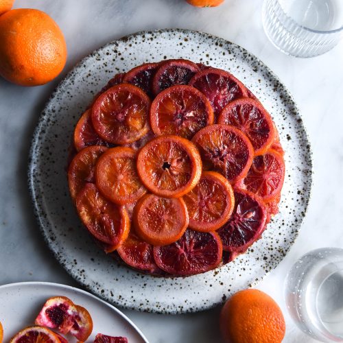 An aerial view of a gluten free upside down blood orange cake. The cake sits atop a white speckle ceramic plate on a white marble backdrop. The backdrop is strewn with extra blood oranges, blood orange peel and glasses of water.