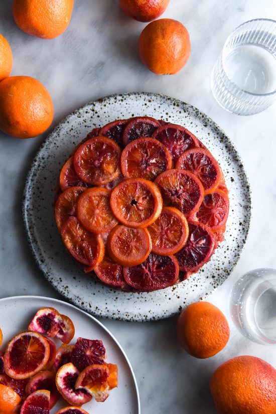 An aerial view of a gluten free upside down blood orange cake. The cake sits atop a white speckle ceramic plate on a white marble backdrop. The backdrop is strewn with extra blood oranges, blood orange peel and glasses of water.