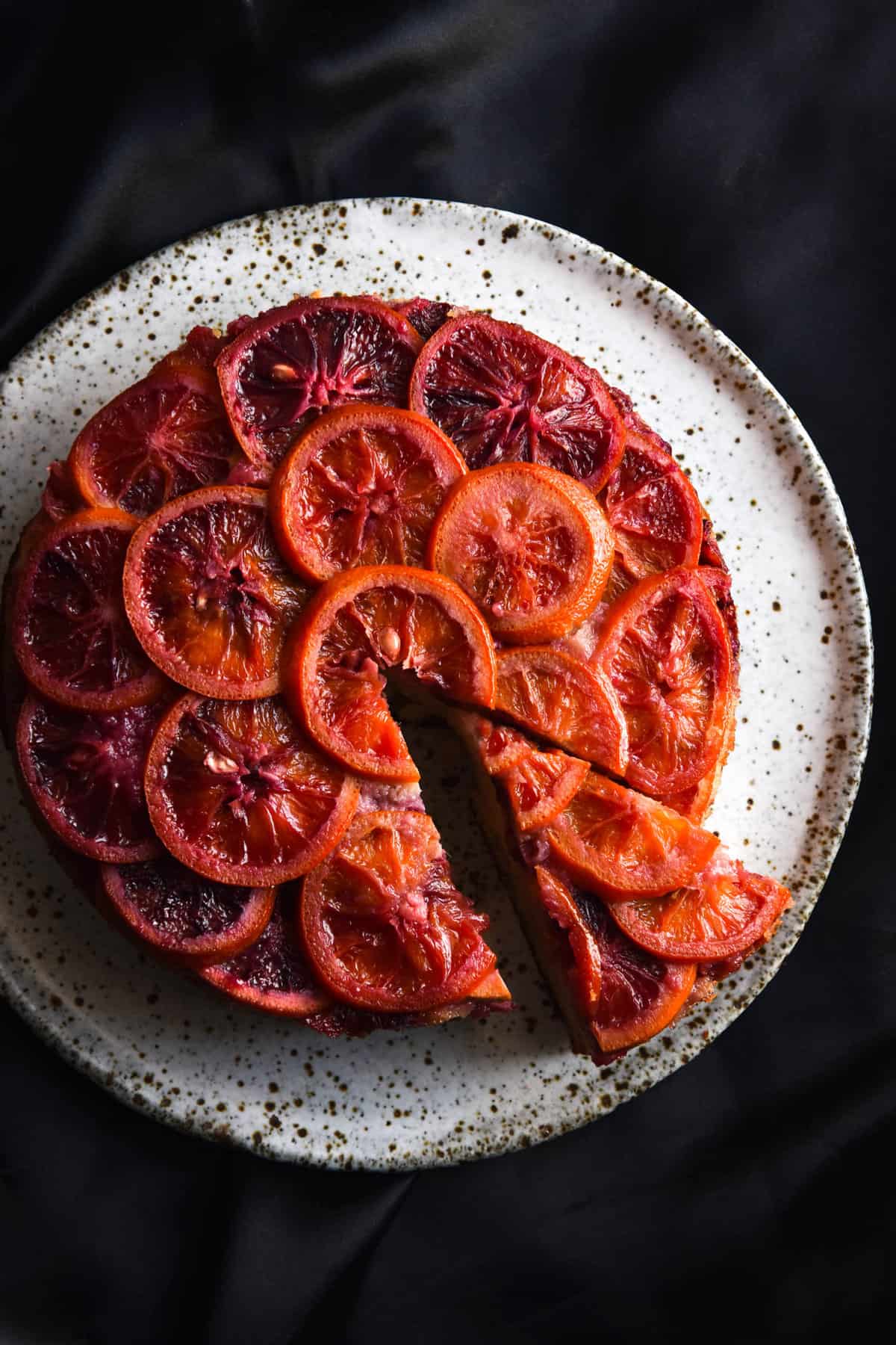 An aerial view of a gluten free upside down orange cake on a speckled white ceramic plate. The plate sits atop a black silky backdrop which contrasts beautifully with the hombre red and orange hues of the blood oranges atop the cake