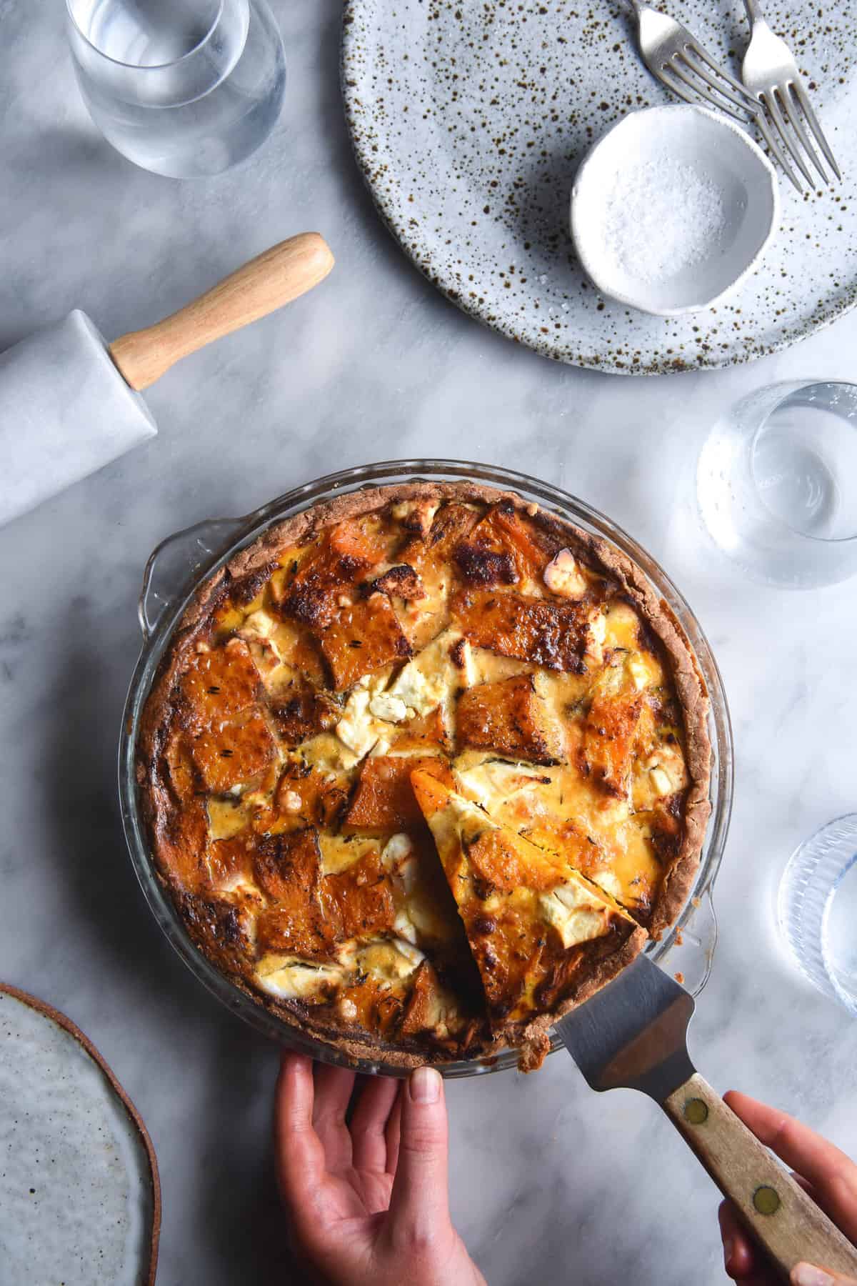 An aerial image of a gluten free pumpkin, sage and goat's cheese tart atop a white marble table. The tart is surrounded by plates, glasses and a marble rolling pin. Two hands extend from the bottom right corner to take a piece of pie.