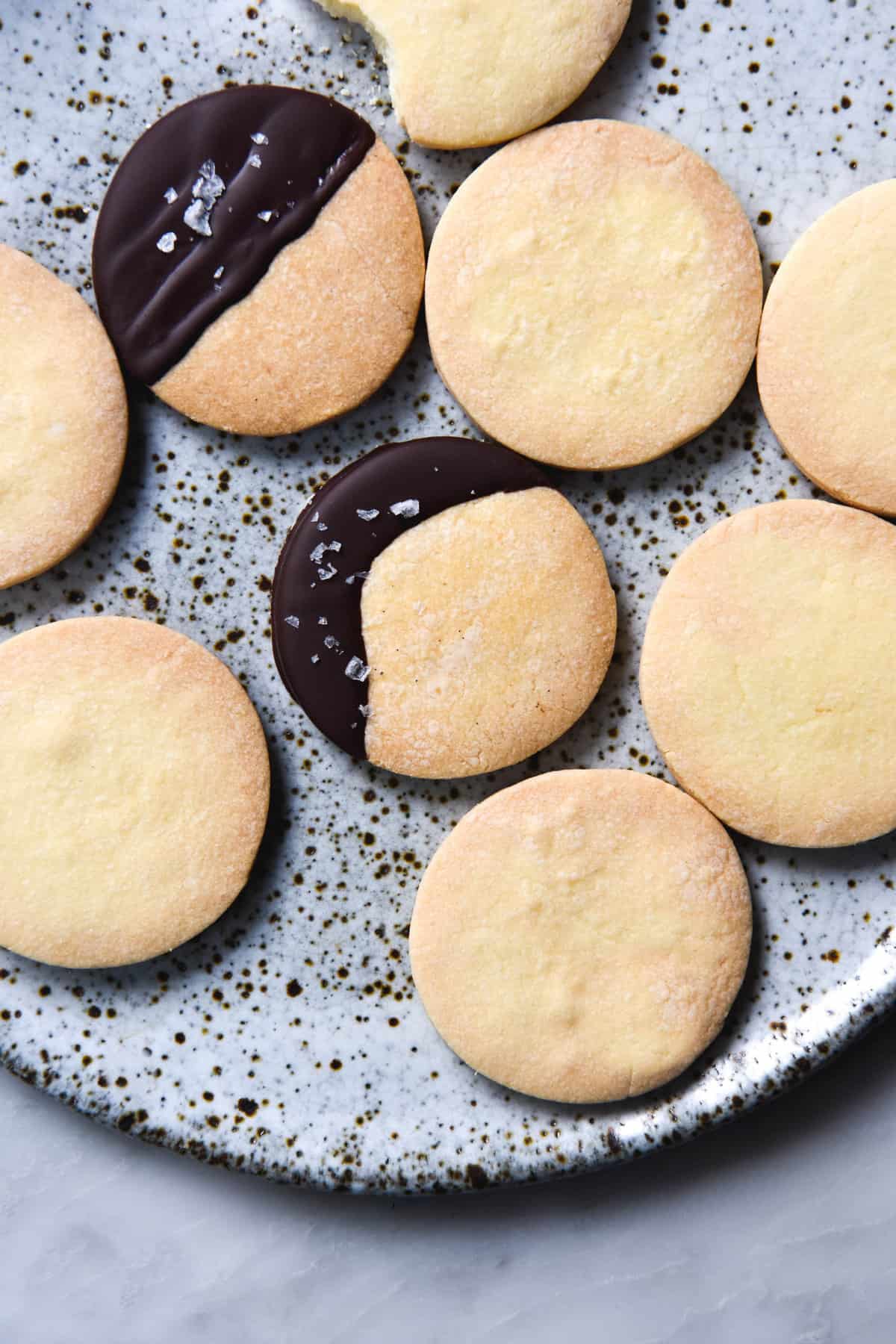 An aerial close up image of gluten free shortbreads on a white speckled ceramic plate. Two of the shortbreads have been dipped in chocolate and sprinkled with sea salt flakes.