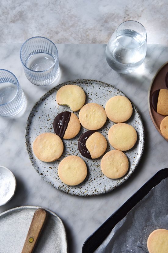 Gluten free shortbreads on a white speckled plate set against a white marble backdrop. Two of the shortbreads are dipped in chocolate and sprinkled with some light sea salt flakes. Surrounding the plate are water glasses, a white ceramic plate, a baking tray and another plate of shortbreads