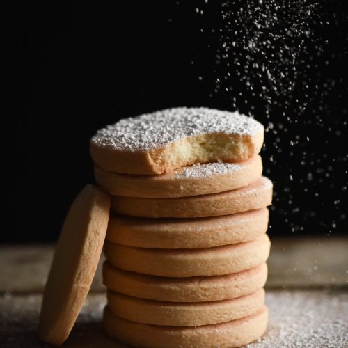 A side on shot of a stack of gluten free shortbread. The shortbread are being dusted with icing sugar from the top right of the image. A single shortbread leans against the stack to the left of the image.