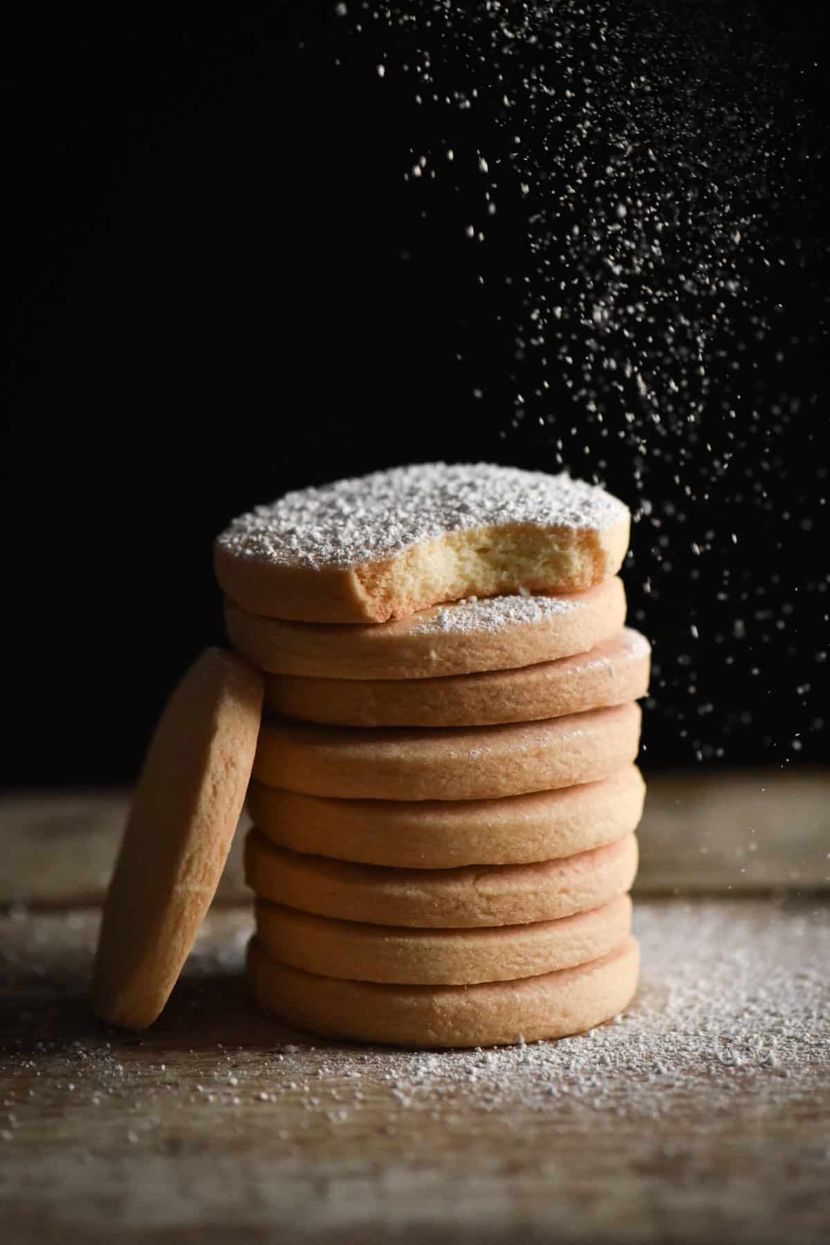 A side on shot of a stack of gluten free shortbread. The shortbread are being dusted with icing sugar from the top right of the image. A single shortbread leans against the stack to the left of the image.