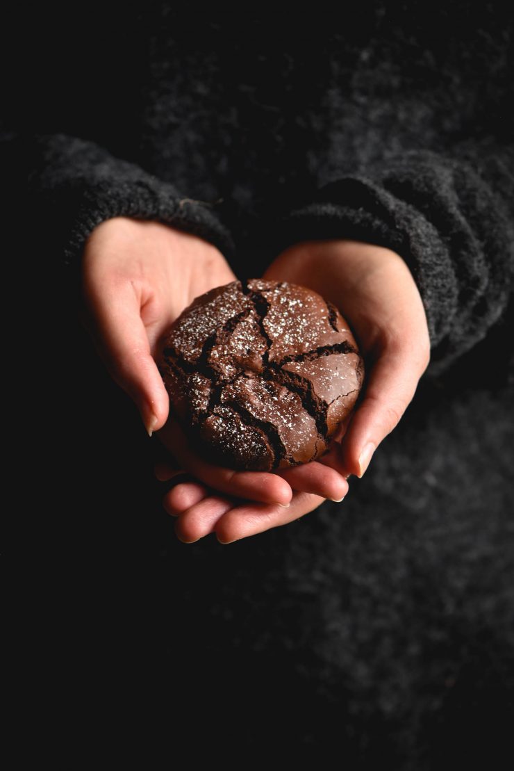 A gluten free chocolate crinkle cookie being held in the palms of female hands. The backdrop is a moody dark woolen jumper and the hands are the central focus of the image. The cookie is crinkled and has been lightly dusted with icing sugar.
