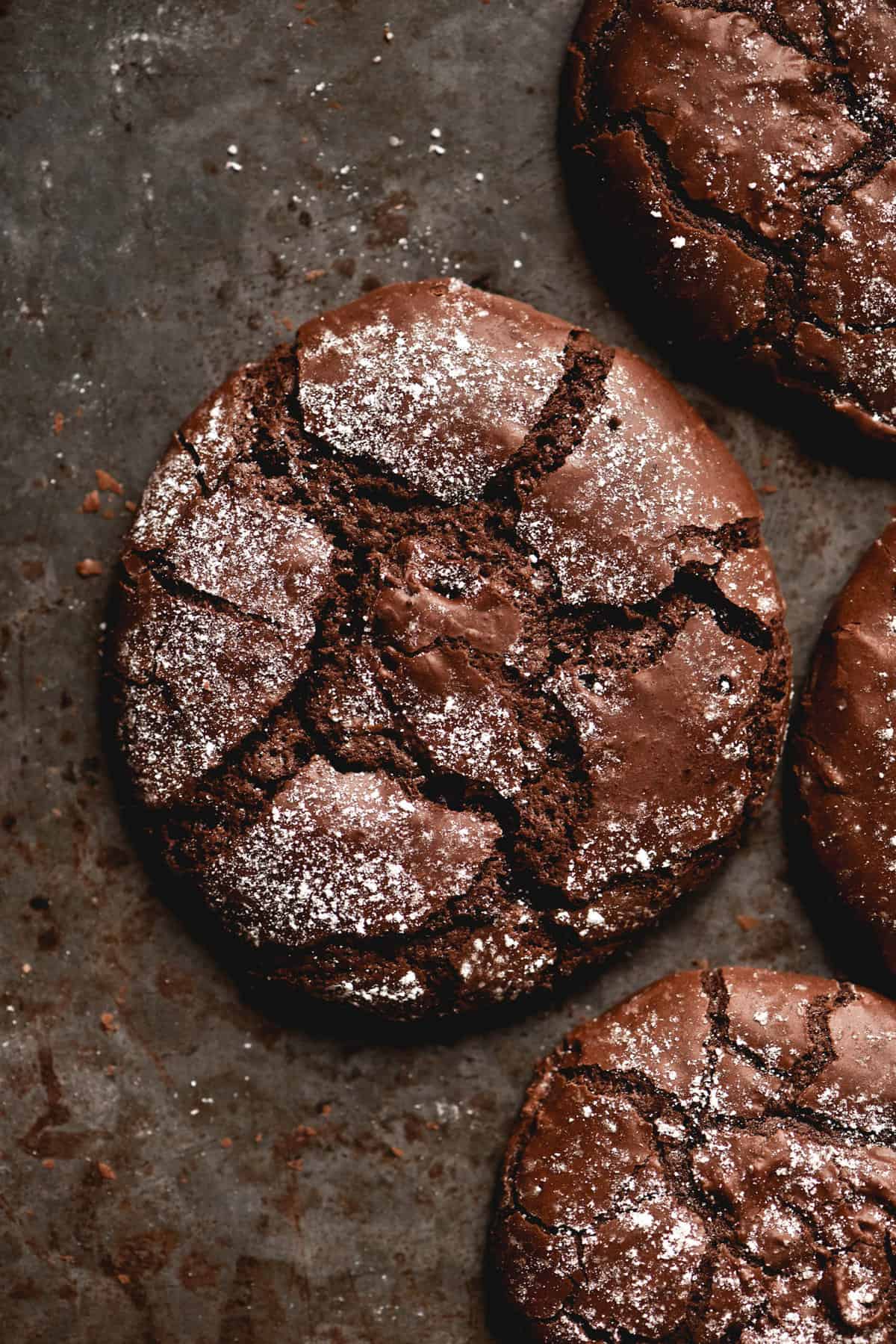 An aerial view of three gluten free chocolate crinkle cookies atop a mottled grey backdrop. The crinkle cookies are a deep chocolate colour and sprinkled with icing sugar, of which flecks are seen against the dark backdrop