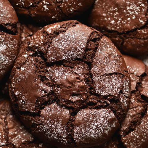 A close up aerial shot of a plate of gluten free chocolate crinkle cookies. The cookies sit in a pile atop a white speckled ceramic plate. They are a deep chocolate colour which contrasts with the icing sugar dusted across their surface.