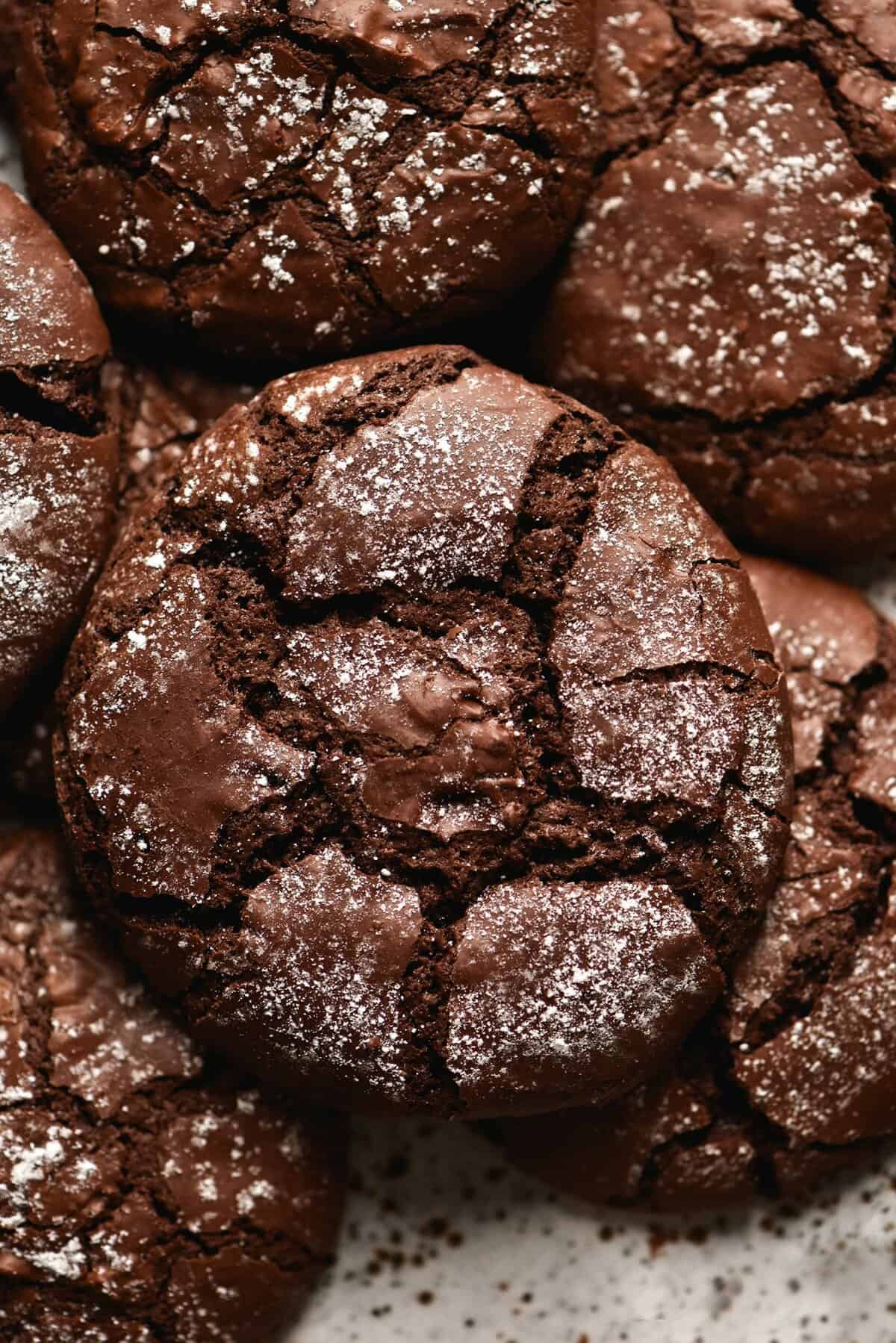 A close up aerial shot of a plate of gluten free chocolate crinkle cookies. The cookies sit in a pile atop a white speckled ceramic plate. They are a deep chocolate colour which contrasts with the icing sugar dusted across their surface.
