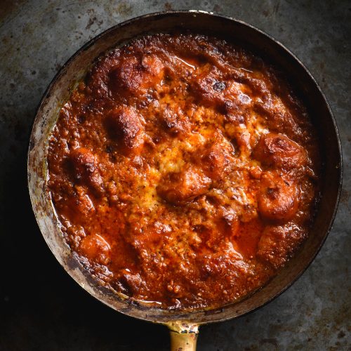 A small skillet of gluten free potato gnocchi baked in a rich FODMAP friendly pasta sauce and topped with melted cheese. The skillet sits against a dark grey metal backdrop.