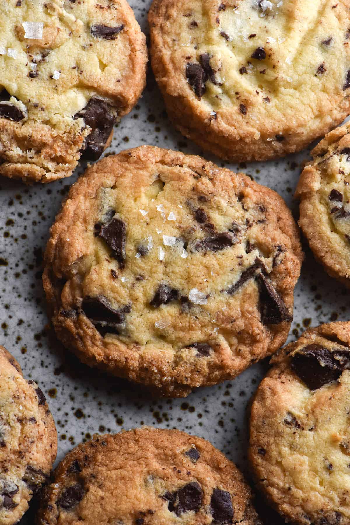 An aerial close up view of gluten free choc chip shortbread on a white speckled ceramic plate. The shortbread are golden brown and topped with a sprinkle of sea salt flakes