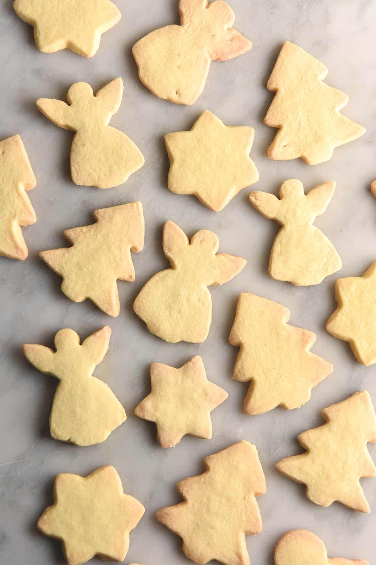 An aerial view of gluten free shortbread cut into various festive shapes - Christmas trees, angels and stars. The shortbread sit atop a white marble table.