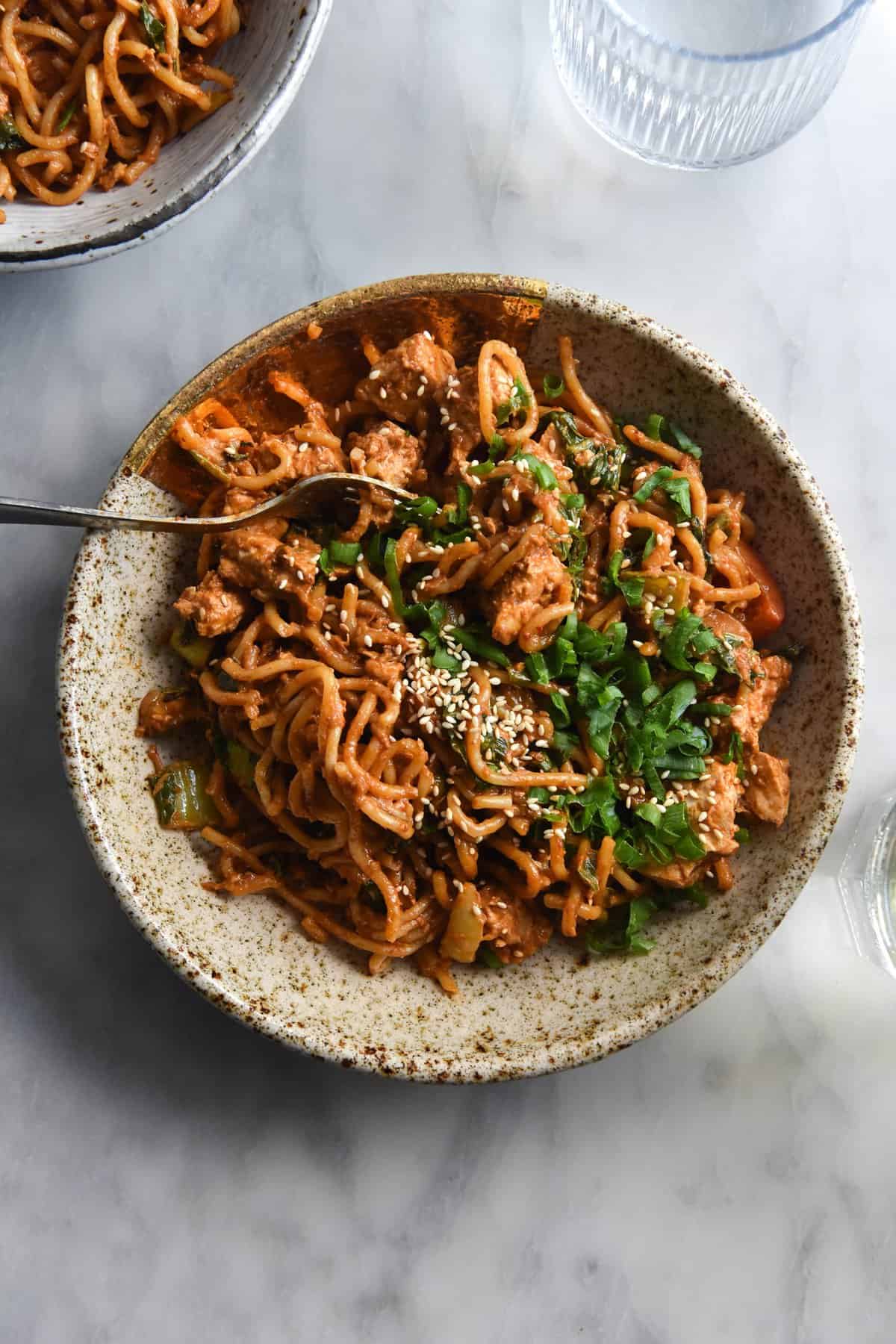 An aerial view of gluten free peanut butter noodles in a speckled ceramic bowl. The noodles are topped with spring onion greens and toasted sesame seeds. They sit atop a white marble table and a second bowl of noodles sits in the top left hand corner