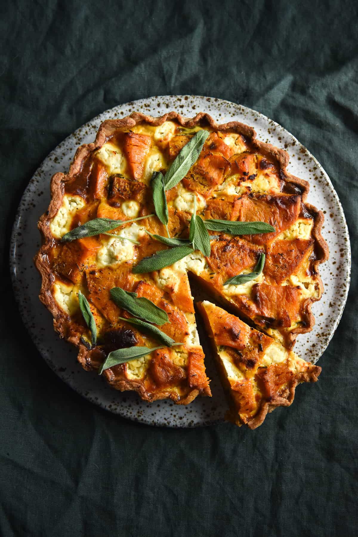 An aerial image of an open topped pumpkin, sage and goat's cheese tart on a white ceramic plate atop an olive green linen backdrop