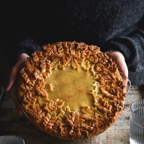 A side on view of a gluten free pumpkin, sage and goats cheese tart with an ornamental pie lid. The pie is covered with pastry and surrounded by a thick border of pastry leaves that have become golden brown in the oven. The pie is being held by two female hands in a wooly grey jumper. A salt dish and a glass of water sit in the foreground on the mottled wooden backdrop