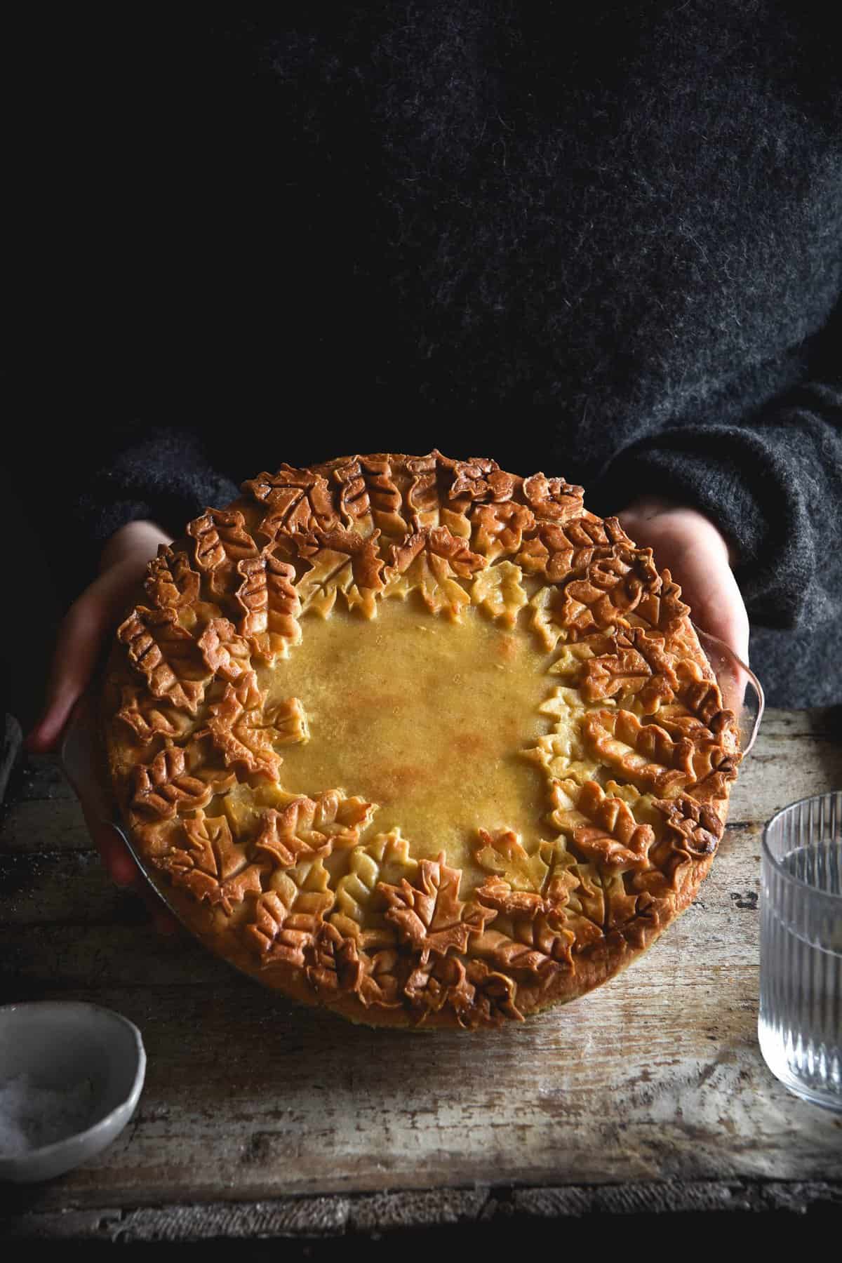 A side on view of a gluten free pumpkin, sage and goats cheese tart with an ornamental pie lid. The pie is covered with pastry and surrounded by a thick border of pastry leaves that have become golden brown in the oven. The pie is being held by two female hands in a wooly grey jumper. A salt dish and a glass of water sit in the foreground on the mottled wooden backdrop