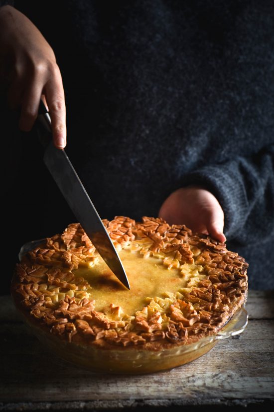 A side on view of a gluten free pumpkin, sage and goats cheese tart with an ornamental pie lid. The pie is covered with pastry and surrounded by a thick border of pastry leaves that have become golden brown in the oven. The pie is being held by two female hands in a wooly grey jumper. A hand extends with a knife to begin cutting into the tart