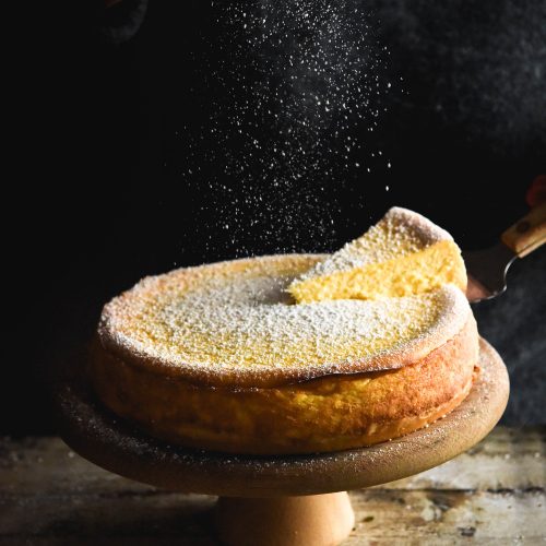 A lactose free, gluten free cheesecake with a shortbread base set against a dark backdrop. It sits on a wooden backdrop and is being sprinkled with a dusting of icing sugar