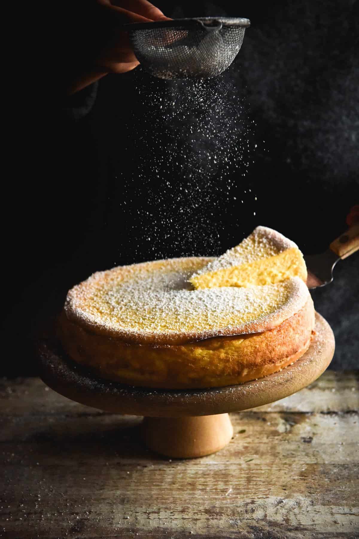 A lactose free, gluten free cheesecake with a shortbread base set against a dark backdrop. It sits on a wooden backdrop and is being sprinkled with a dusting of icing sugar