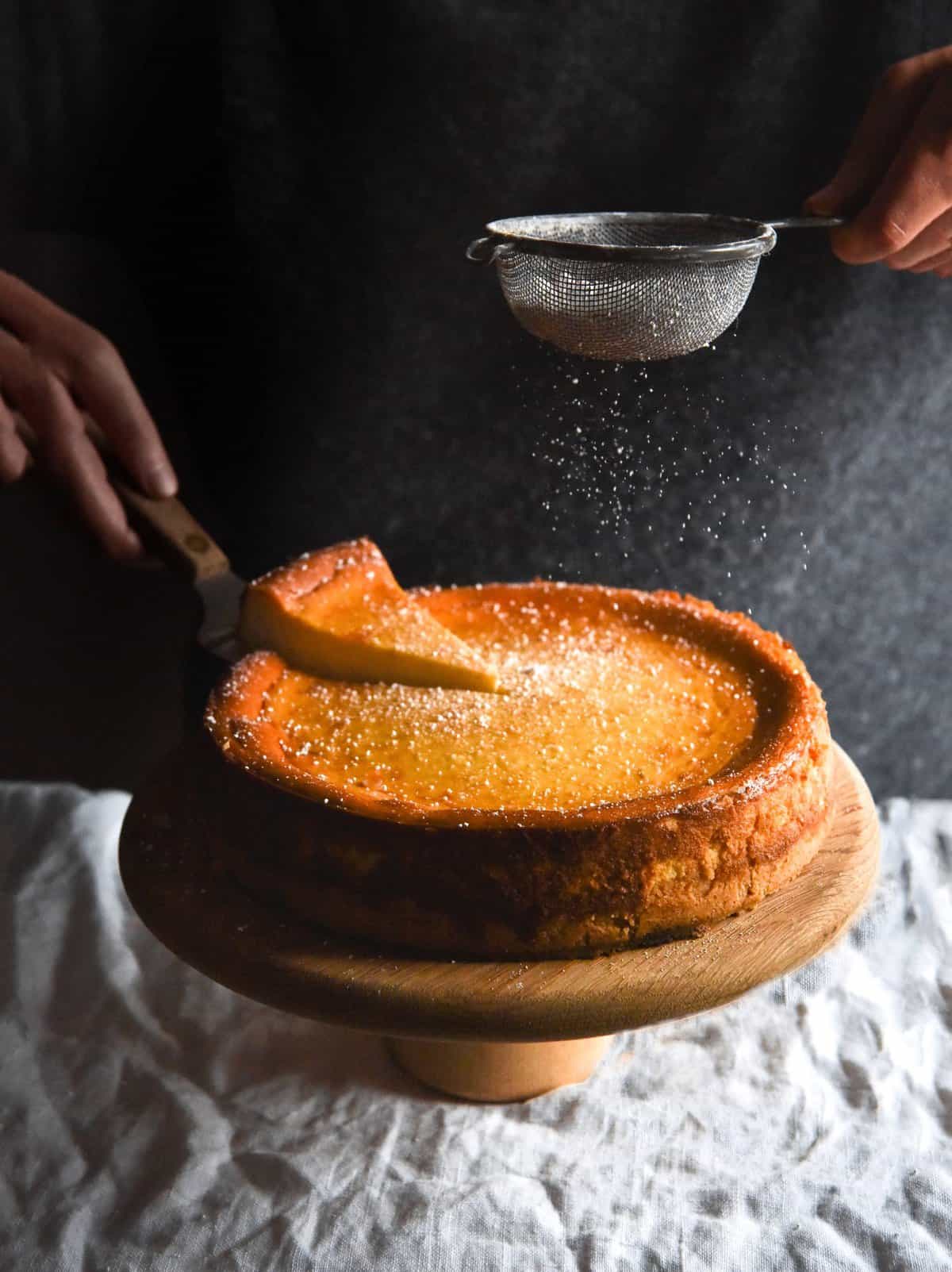 A lactose free, gluten free cheesecake with a shortbread base set against a dark backdrop. It sits on a wooden backdrop and is being sprinkled with a dusting of icing sugar